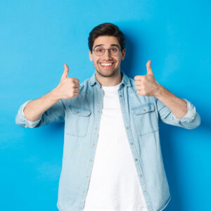 Attractive young man wearing glasses and casual clothes, showing thumbs up in approval, like something, standing against blue background.
