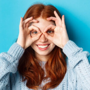 Close-up of funny and cute redhead girl making hand glasses and looking through them, seeing promo offer and smiling, standing over blue background.