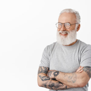 Close up of handsome senior guy with tattoos, wearing glasses, looking left and smiling happy at camera, chatting with someone, standing in grey t-shirt against white background.