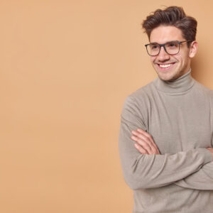 Positive self confident man keeps arms folded looks away with satisfied expression listens good news wears spectaces and casual turtleneck isolated over beige background blank space for promo