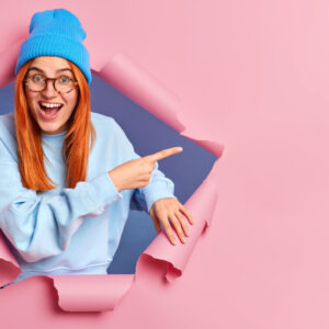 Glad red haired woman suggests to check this promo indicates aside on copy space poses through paper background breaks in hole wears casual blue clothes gives direction to shop with big sales